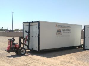 A large white storage container labeled "A Box Moving & Storage" is ready for HOME MOVING on a gravel lot, hitched to a red mover vehicle.