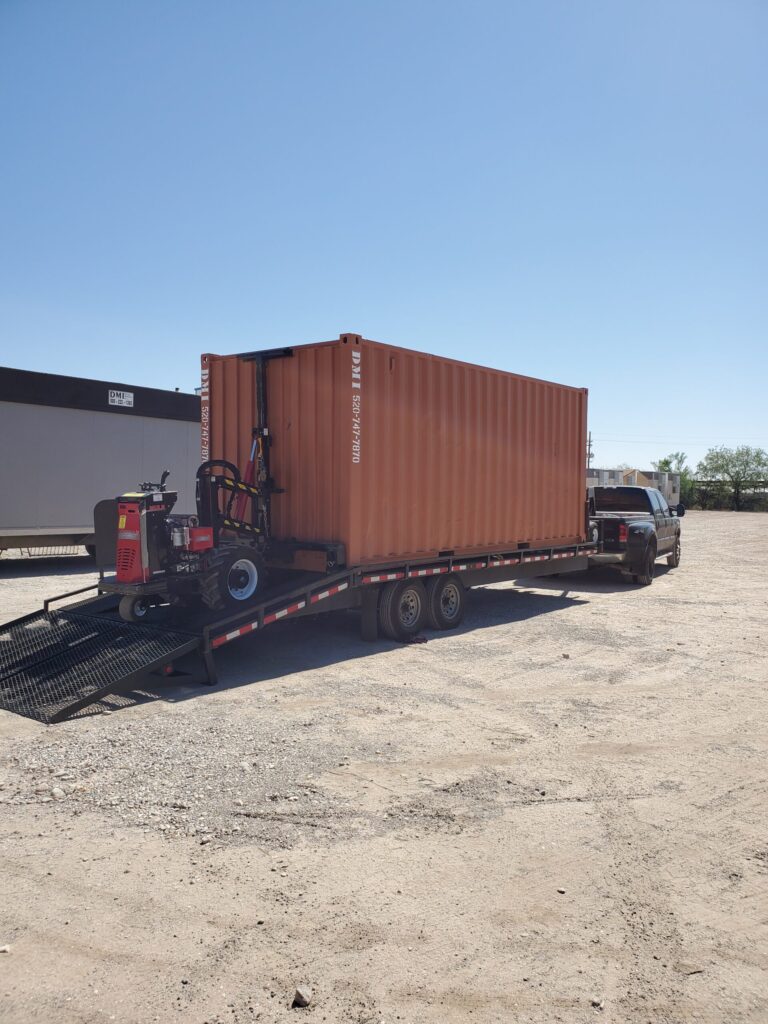 Truck towing a trailer with a large orange shipping container and a red forklift nearby.