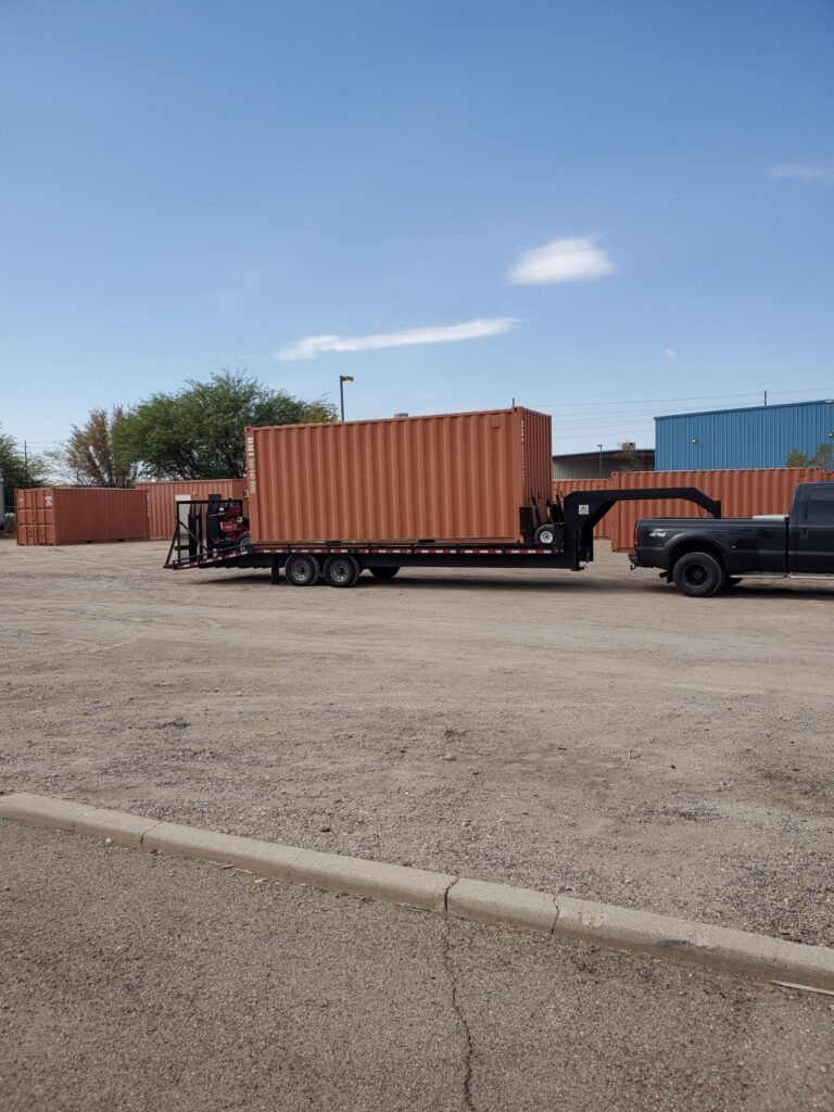 A flatbed trailer carrying a large orange shipping container, attached to a black truck, in a parking area.