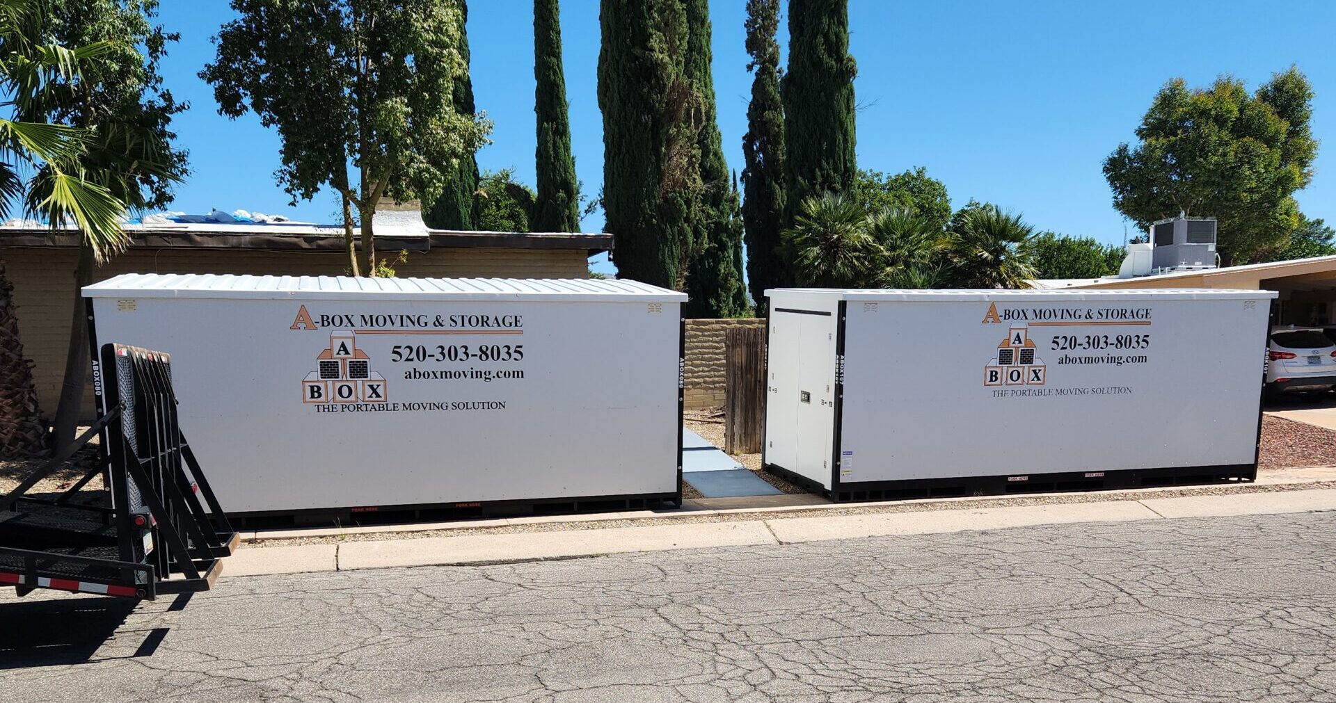 Two white portable storage containers from A BOX Moving & Storage are placed on a residential street next to tall trees.